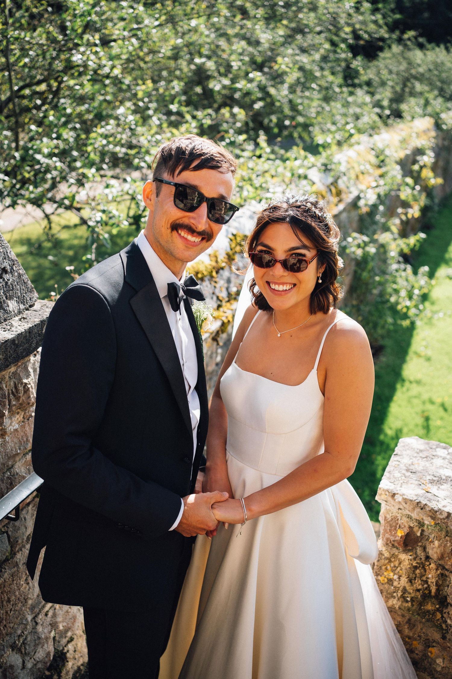 Stylish couple in formal attire share laughs while wearing sunglasses in a garden setting.