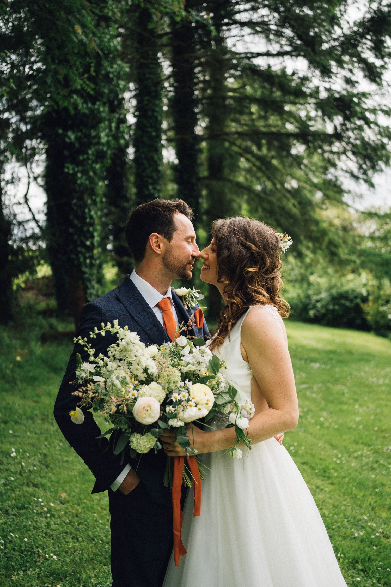 Romantic wedding couple embracing in lush green garden setting with white floral bouquet.