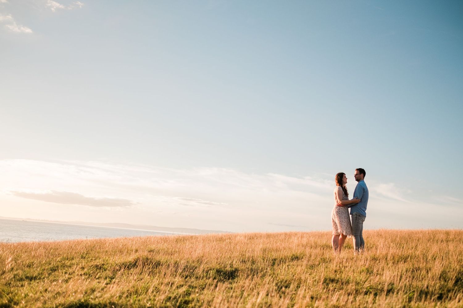 Romantic couple standing together in tall grass field against blue summer sky.