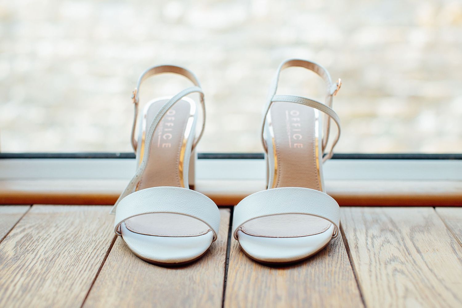 Elegant white bridal sandals displayed on a wooden floor near a window.