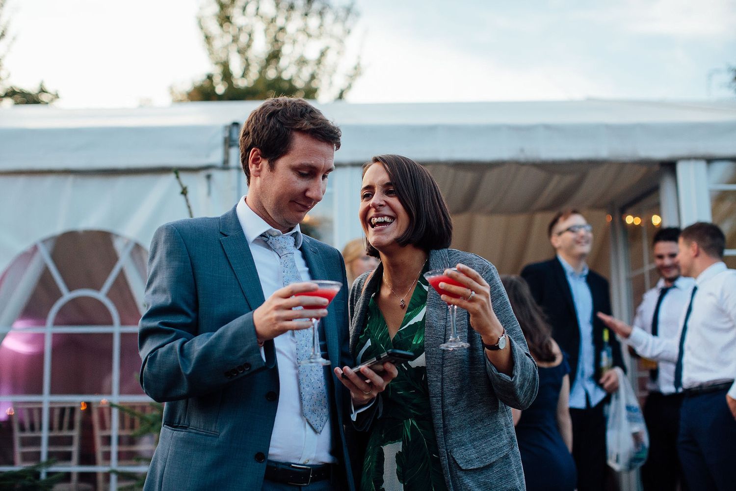 Two guests share a lighthearted moment while enjoying drinks at an elegant outdoor wedding reception.