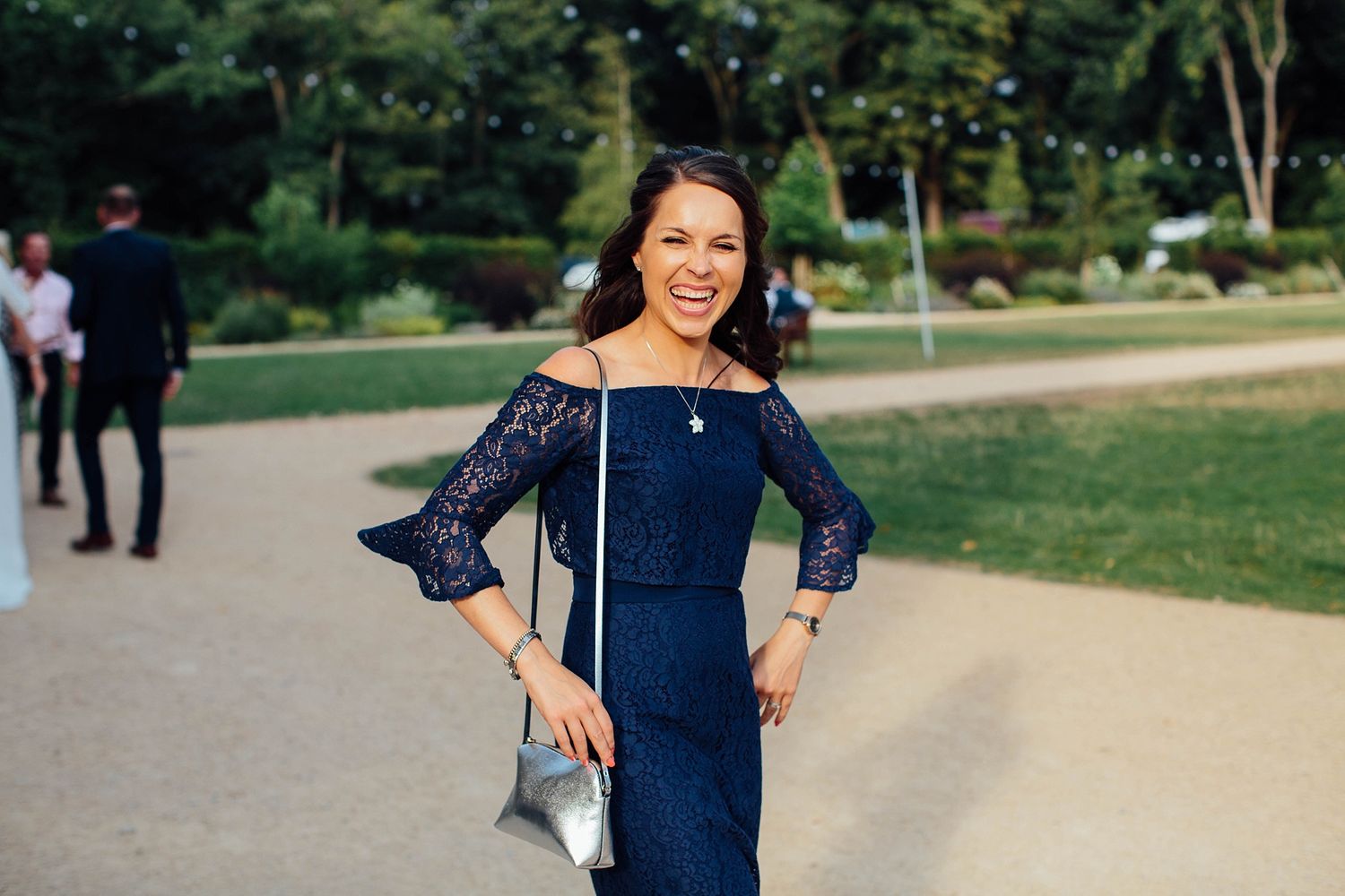 A woman in a navy off-shoulder dress poses on a garden path during a wedding.