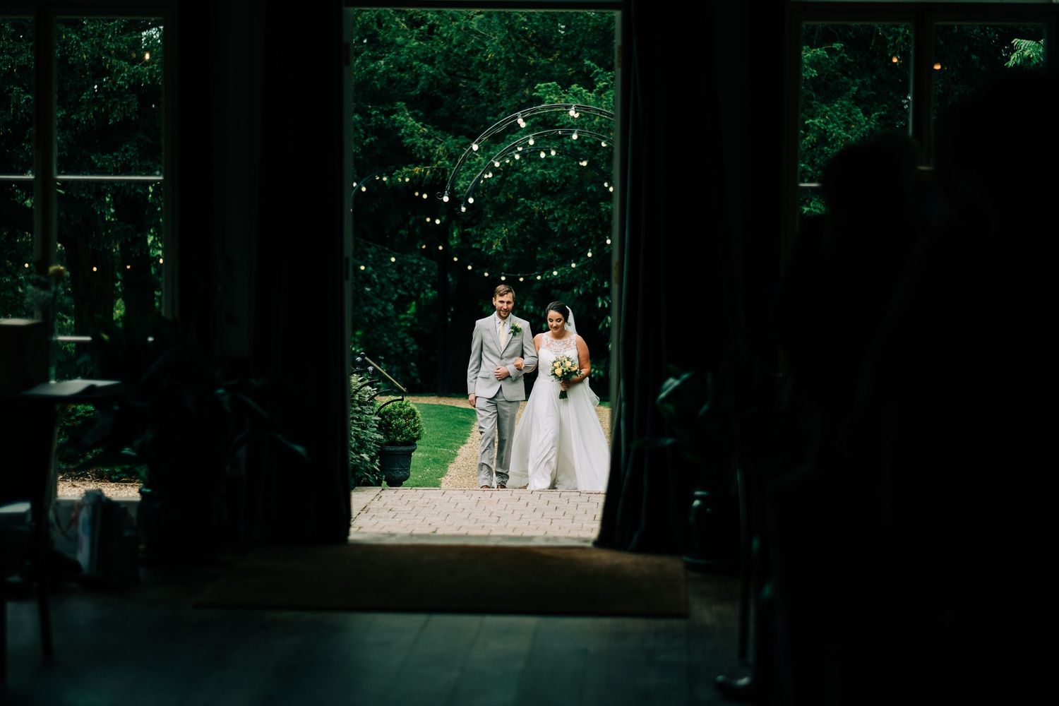 A wedding ceremony scene framed through dark pillars with string lights and greenery.