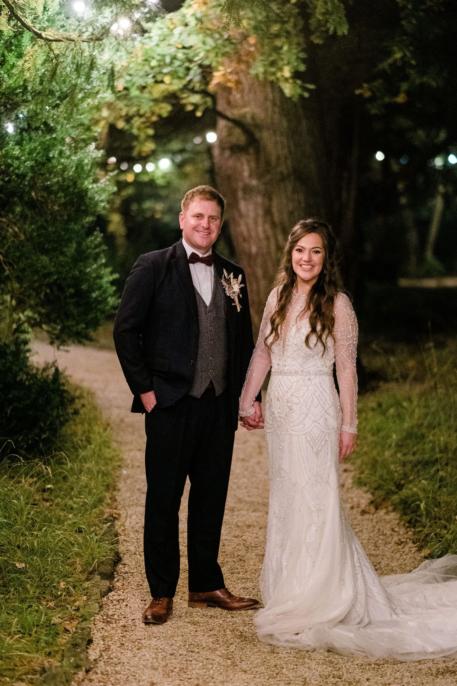 Couple strolls along illuminated garden path during evening wedding photoshoot.