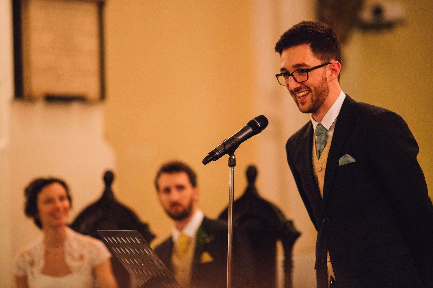A speaker delivers a wedding speech at a reception with guests visible in background.