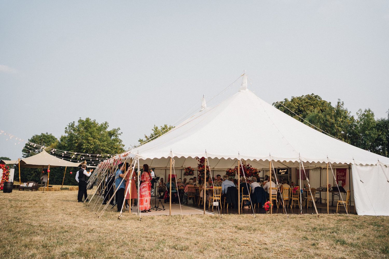 Multiple white wedding reception tents set up on a grassy hillside venue with trees in the background.