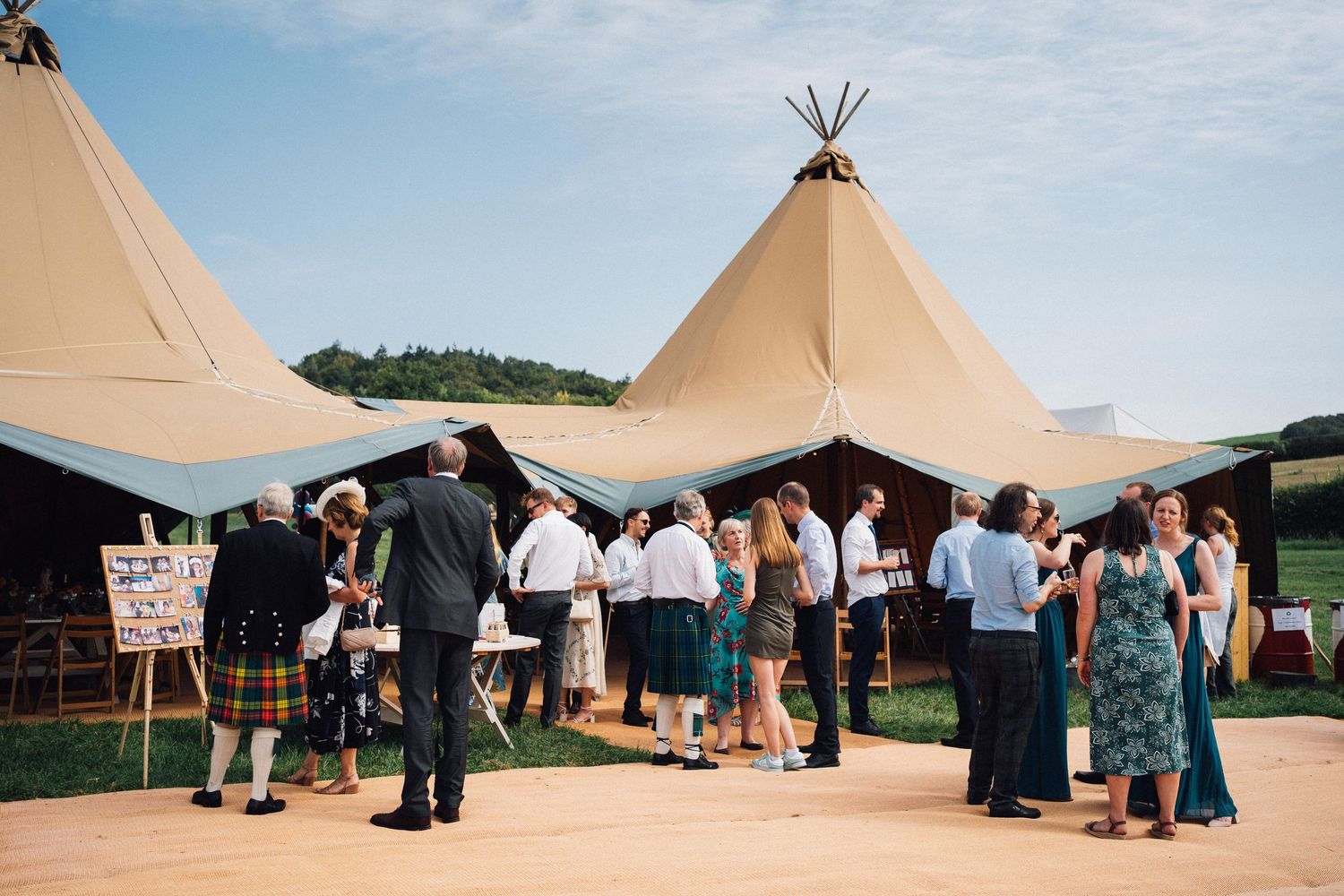 A series of outdoor tipi-style tents set up for an event with guests mingling around them in a rural setting.