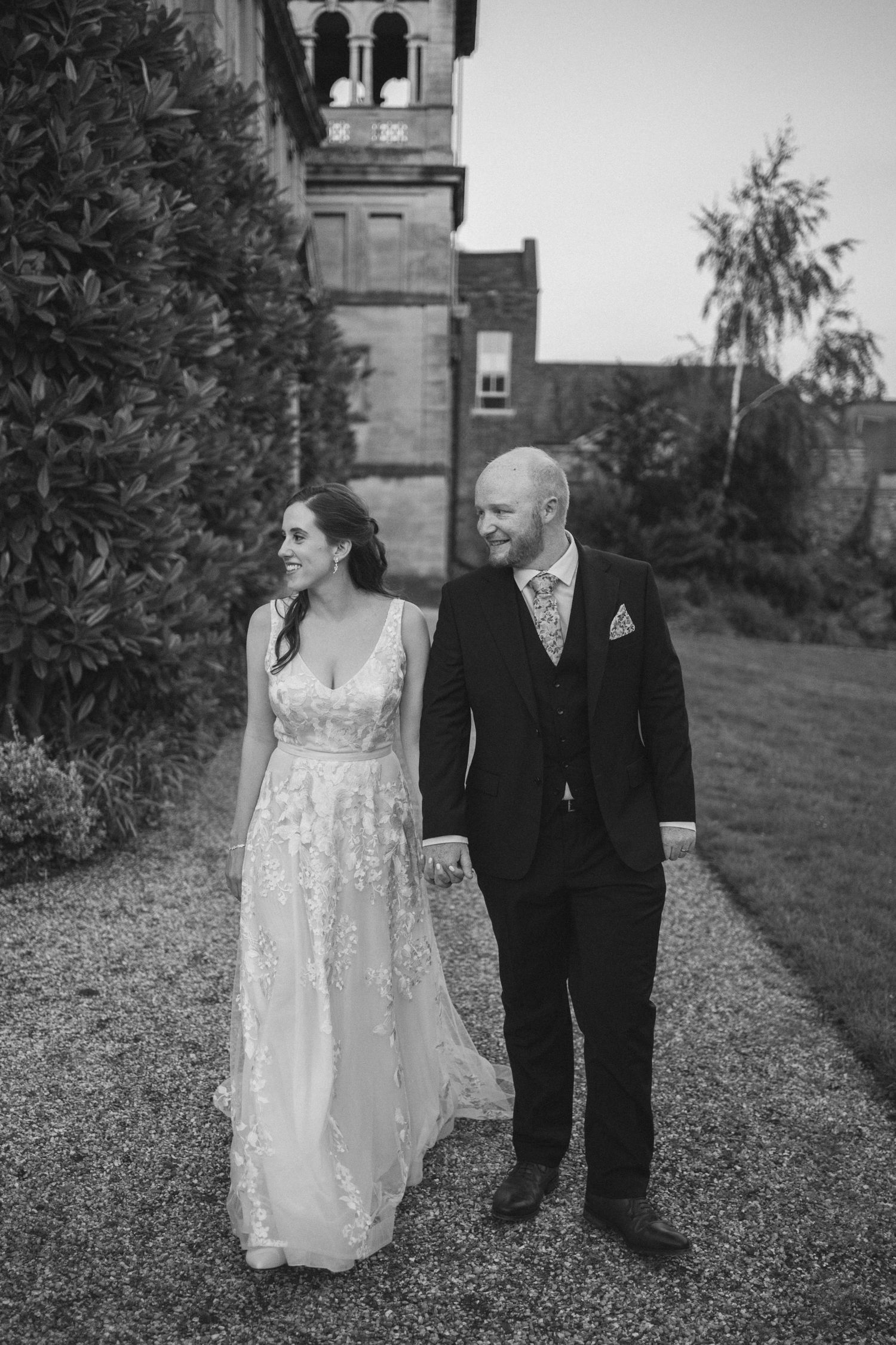A couple in wedding attire walks together on a grassy lawn beside a stone building in black and white photography.