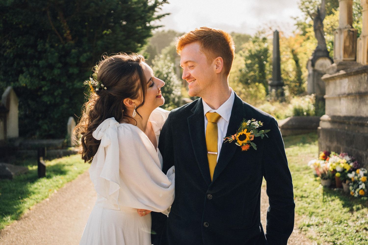 Newlyweds share a tender moment in a sunlit garden path, with the groom wearing a yellow tie and boutonniere.
