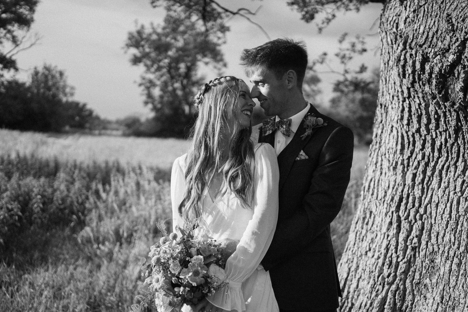 Black and white wedding photo of a couple standing by a tree in a field with the bride holding a wildflower bouquet.