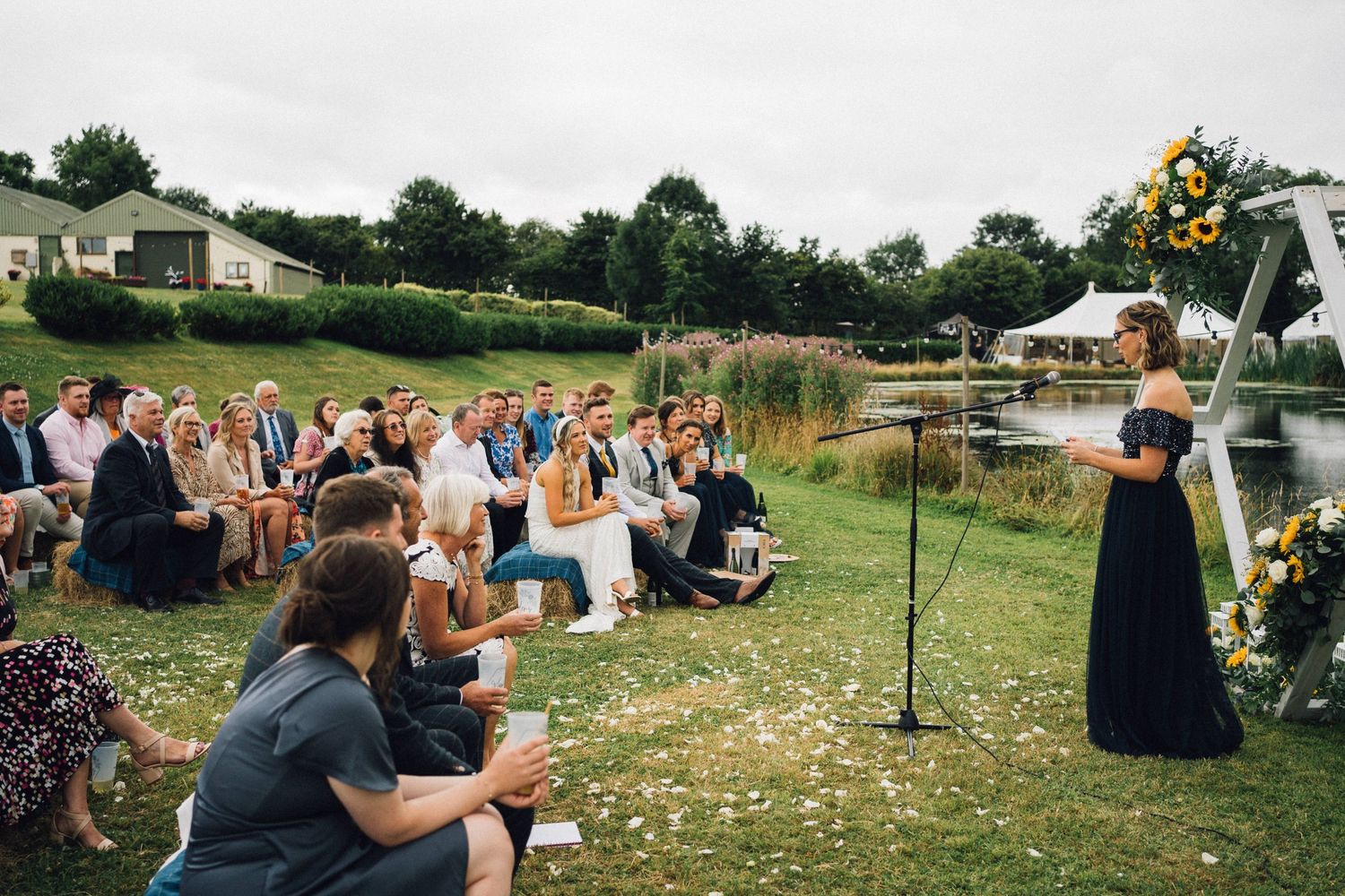 Outdoor wedding ceremony by a pond with guests seated, a woman in black dress speaking at a floral arch.