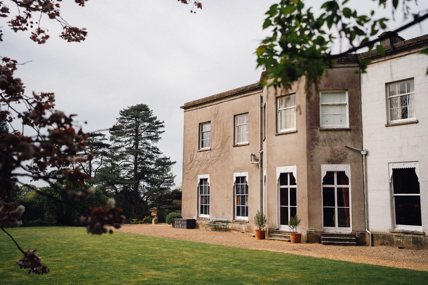 A grand Georgian country house with manicured lawns and tall pine trees under an overcast sky.