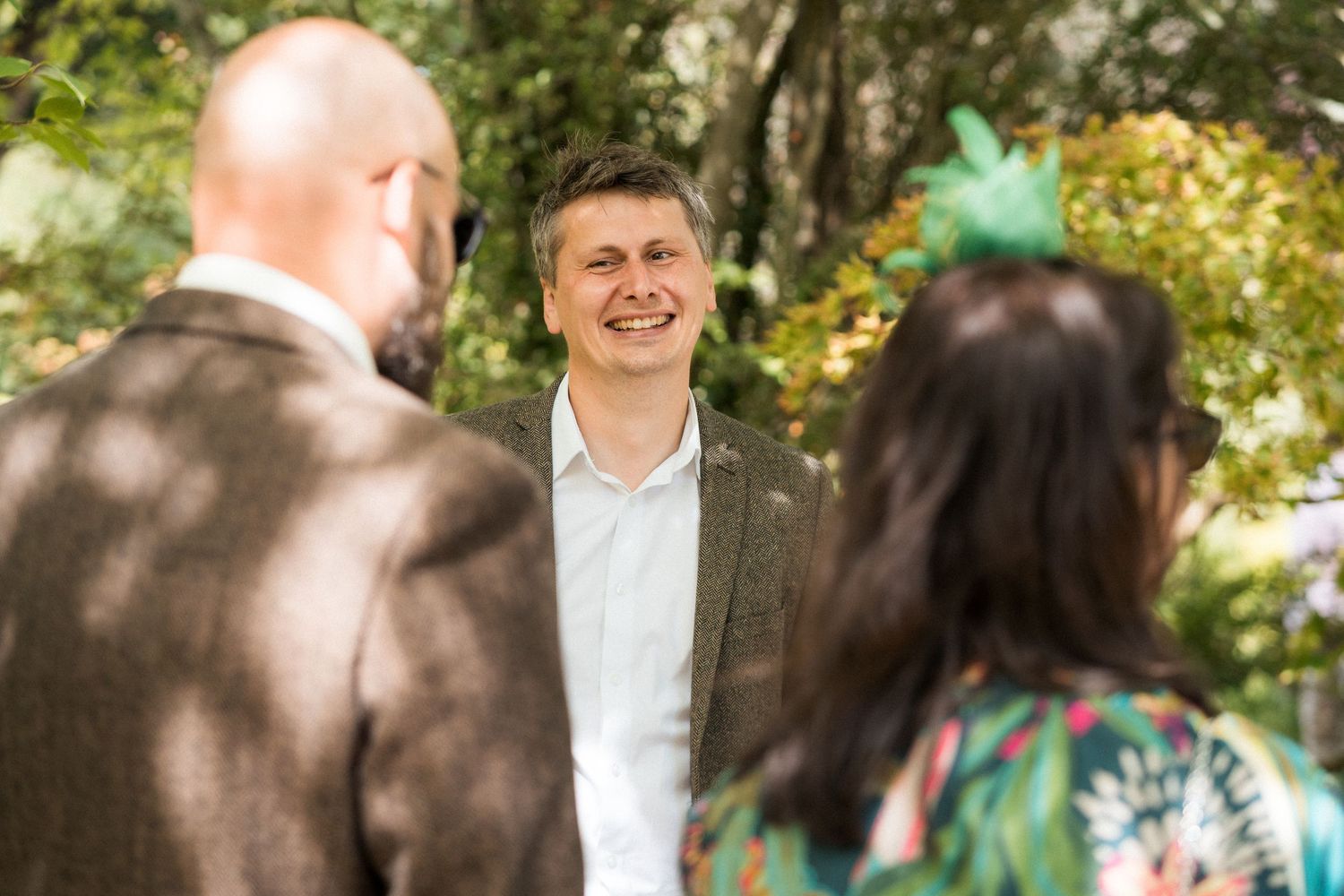 Smiling man in a grey suit jacket conversing with guests outdoors at a garden event surrounded by lush green trees.