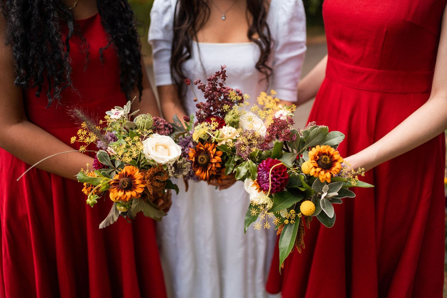 Bride in white gown flanked by bridesmaids in red dresses holding vibrant fall bouquets with sunflowers and seasonal blooms.