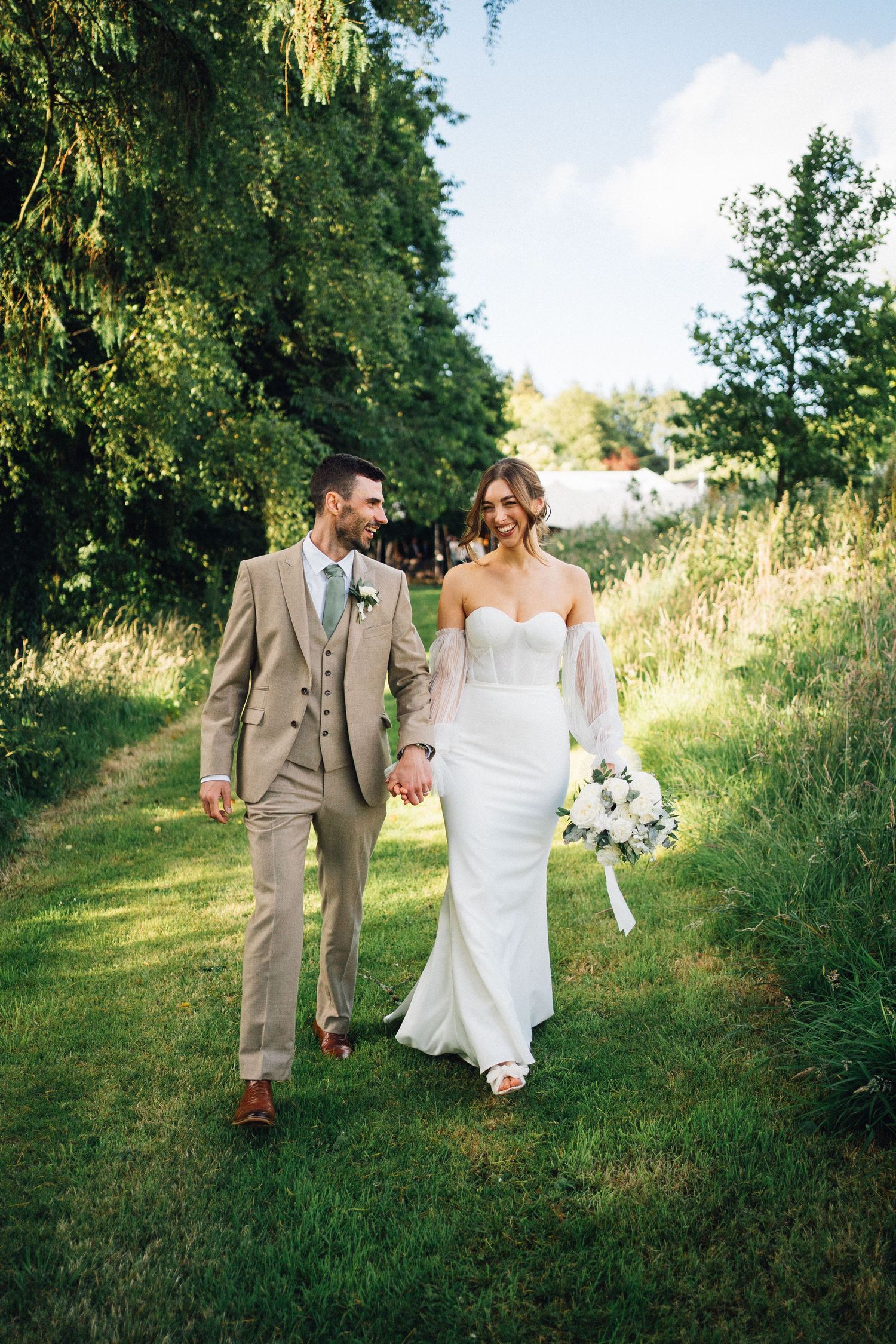 Bride and groom walk hand-in-hand through lush green gardens, she in a white gown, he in a tan suit.