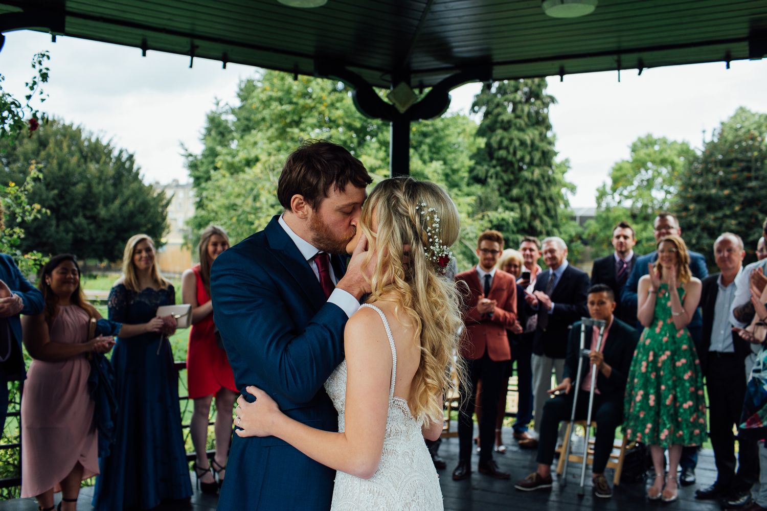 A couple shares their first kiss as newlyweds under a covered pavilion surrounded by wedding guests.