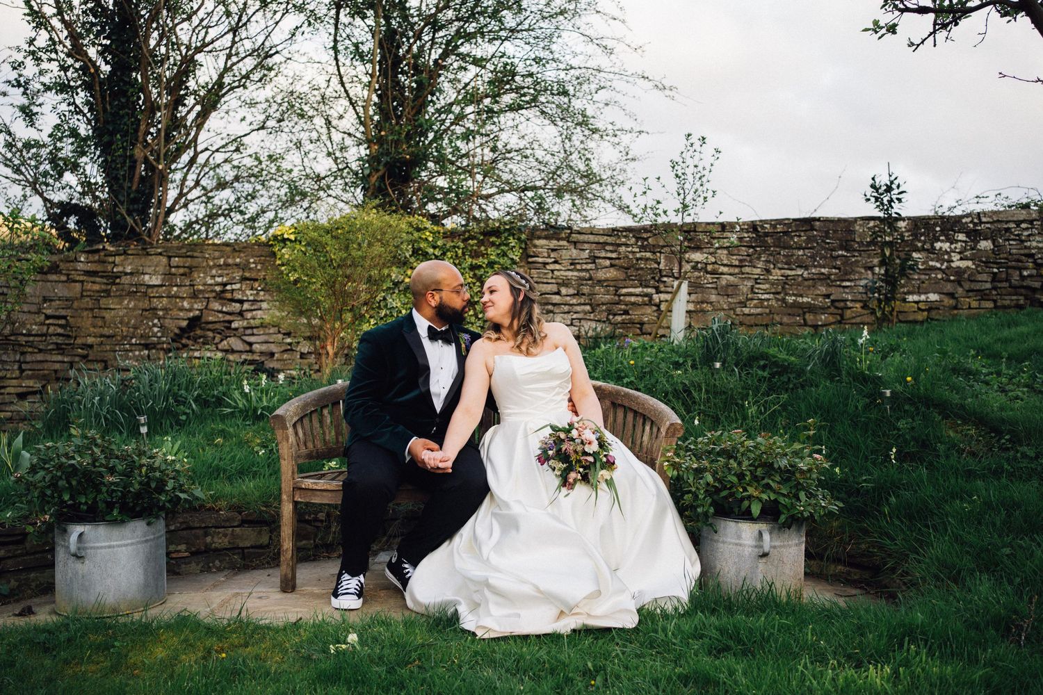 A romantic wedding couple shares intimate moments on a wooden garden bench in an outdoor setting.