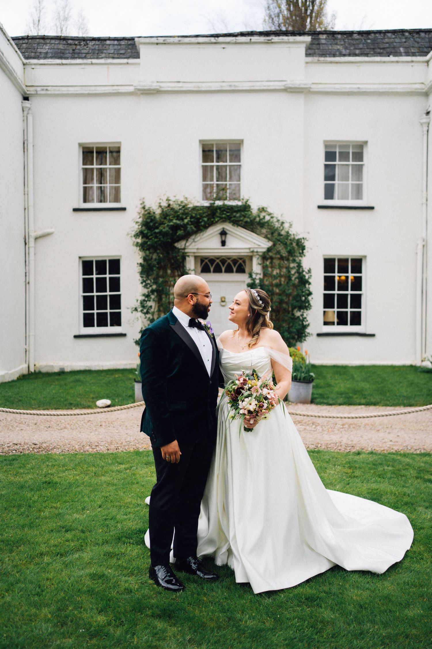 A romantic wedding couple shares intimate moments on a lawn outside a white Georgian-style mansion.