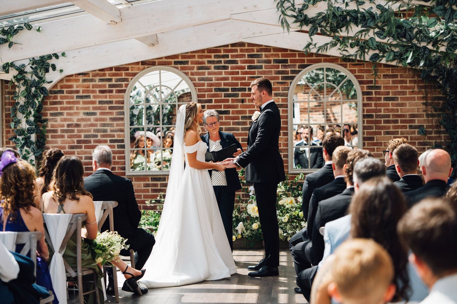 Wedding ceremony taking place under a brick archway with guests seated outdoors on a sunny day.