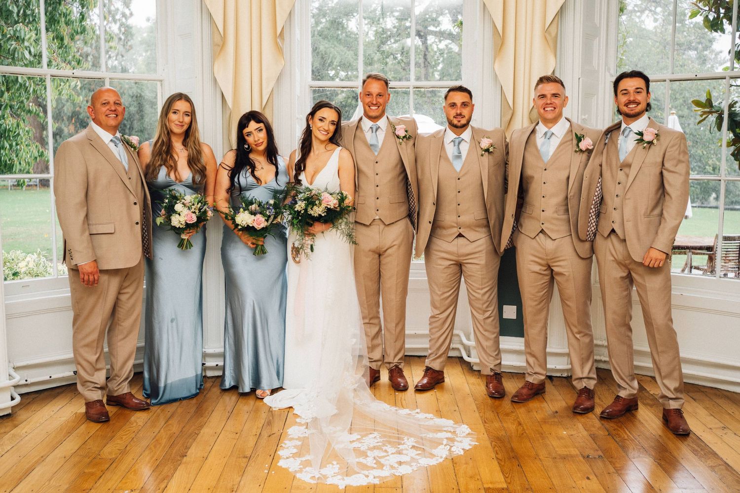 Wedding party in tan suits and light blue dresses posed together in bright indoor venue.