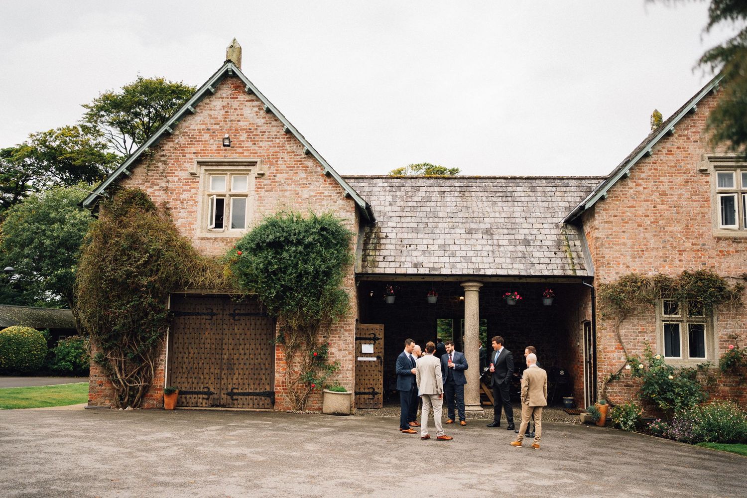 Rustic brick stable building with ivy-covered walls and wooden doors where guests gather at the entrance.