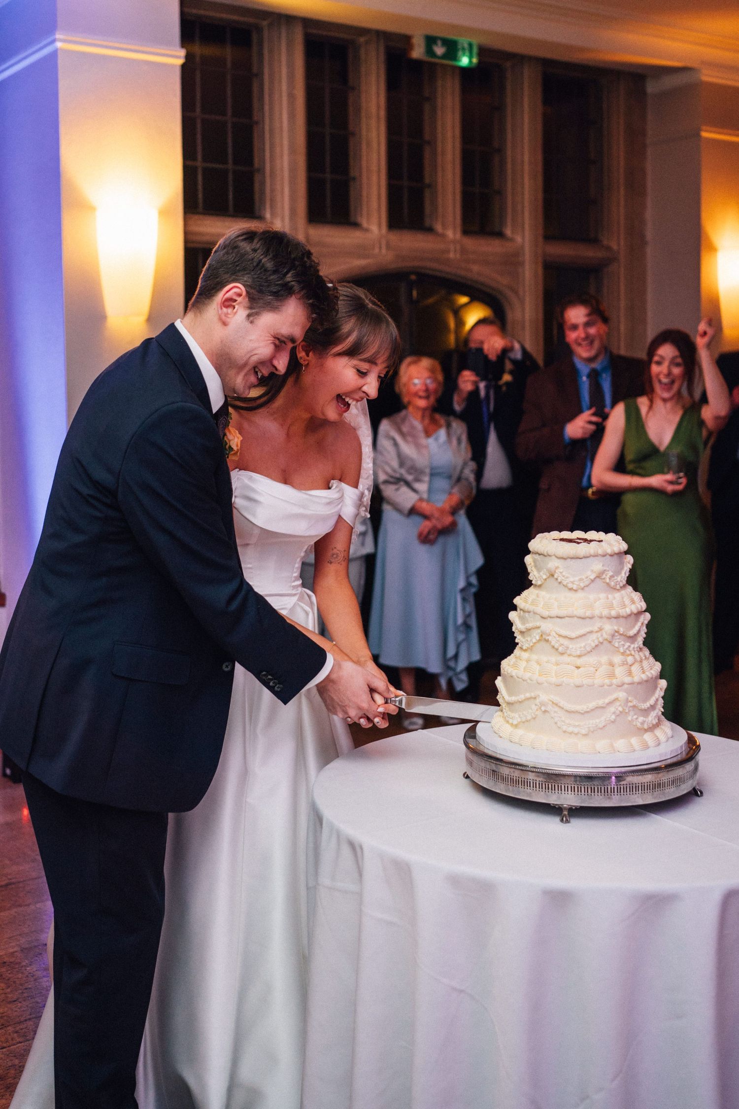 A couple cuts their elegant white tiered wedding cake together while guests watch in the background.