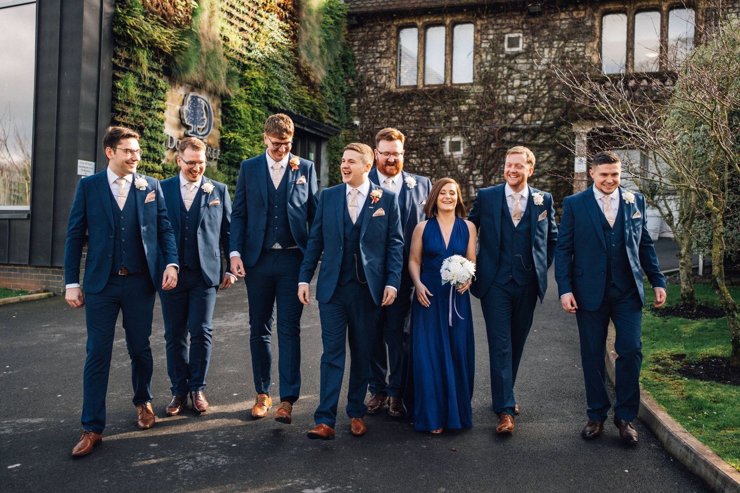 Wedding party members in navy suits walk together in a line along a brick building.