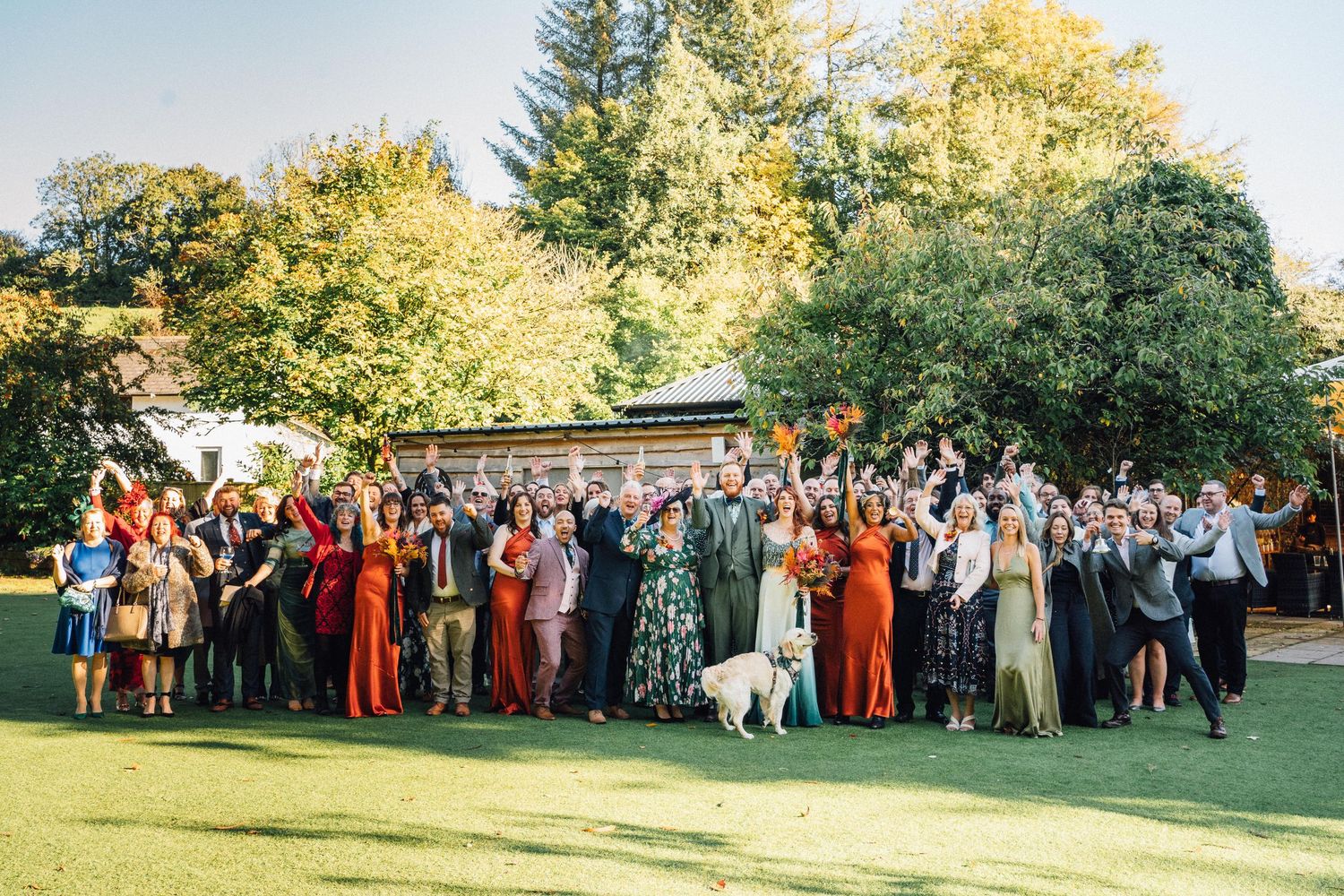 Large group of wedding guests in formal attire gather on sunny lawn for outdoor celebration photo.
