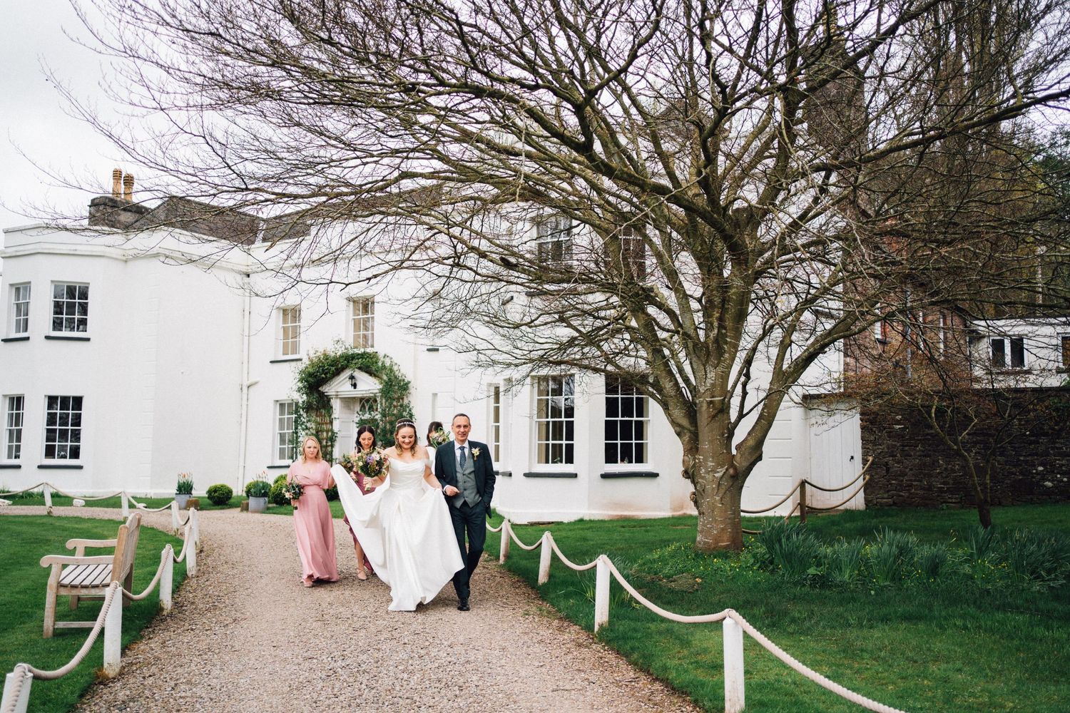 A wedding party walks along a tree-lined gravel path leading to a white historic mansion on a sunny day.