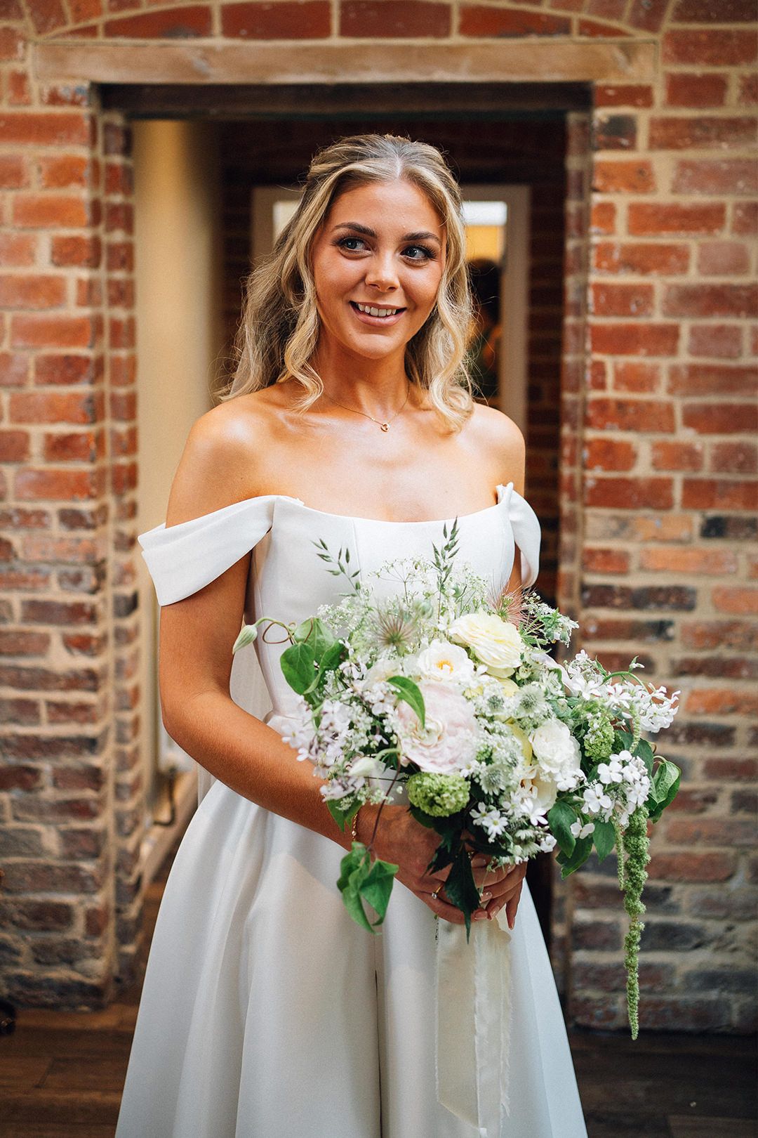 A bride in an off-shoulder white wedding gown holds a lush bouquet of white and green flowers against a brick wall backdrop.