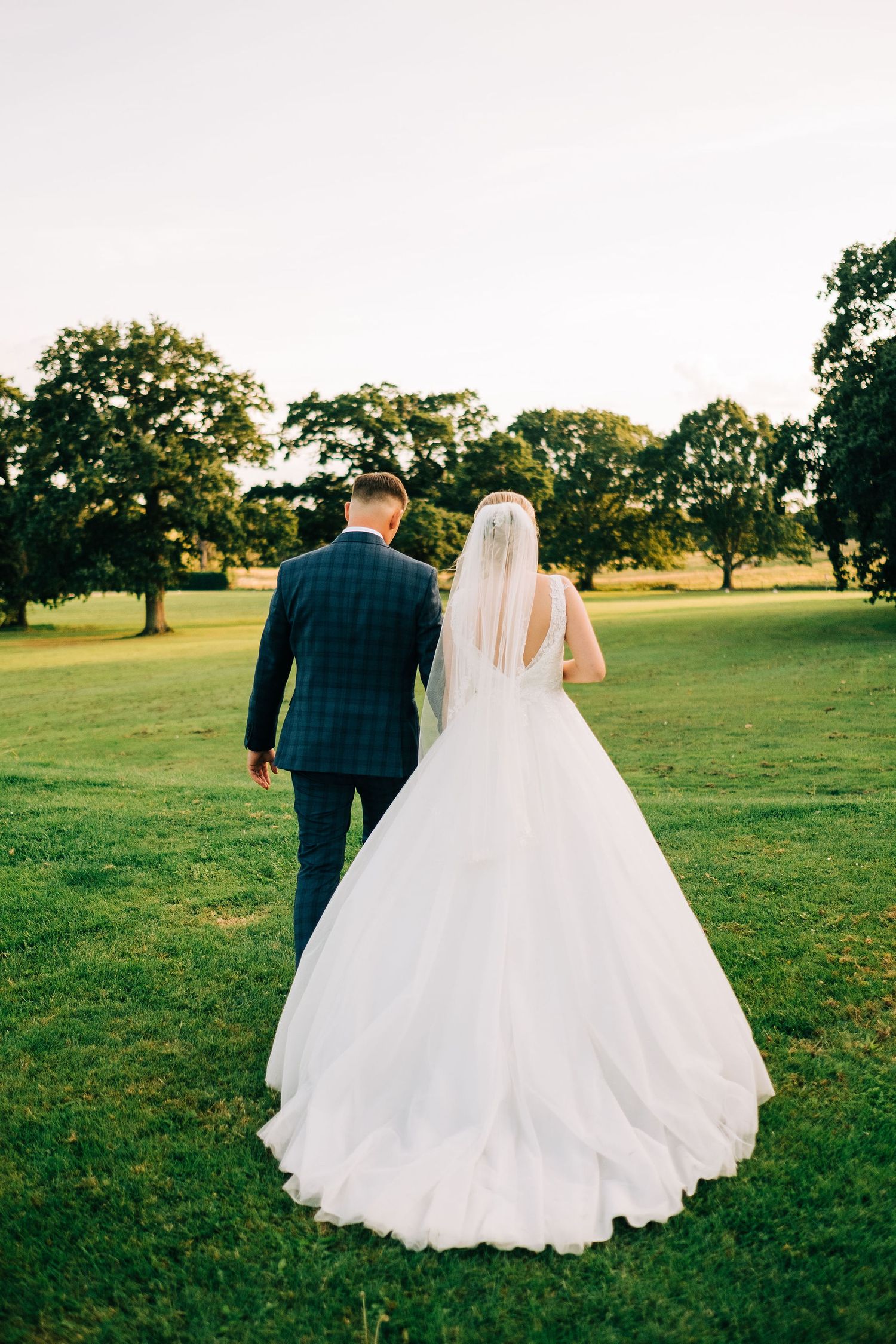 Couple in wedding attire walking across sunlit meadow.
