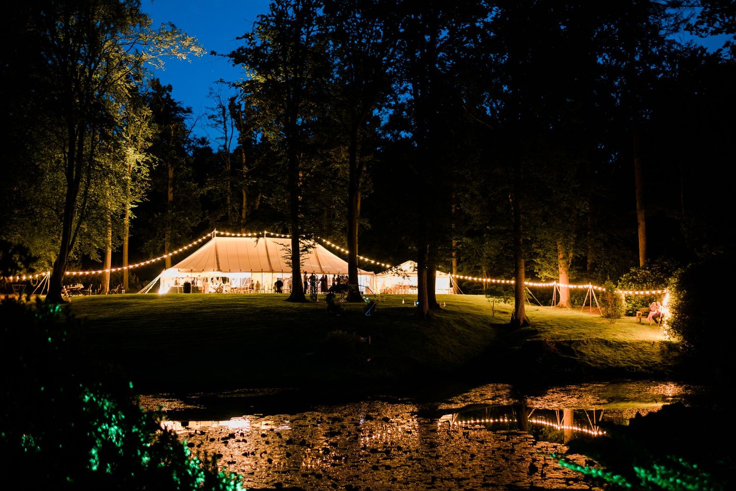 An illuminated wedding tent glows warmly against the dark night sky surrounded by trees.