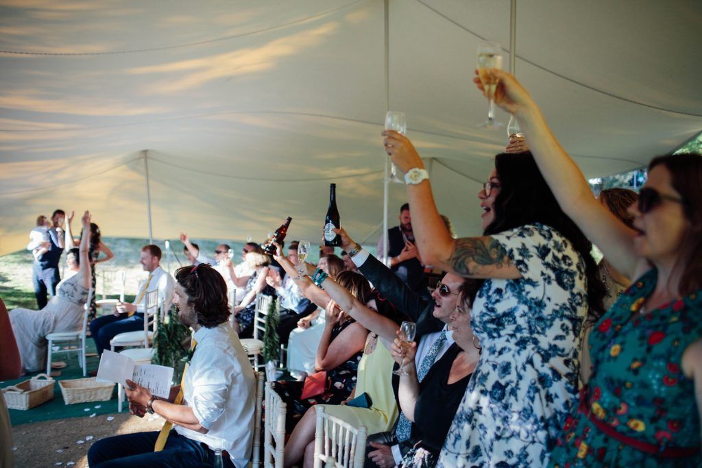 Wedding reception guests raise their glasses in a toast under a white tent canopy.