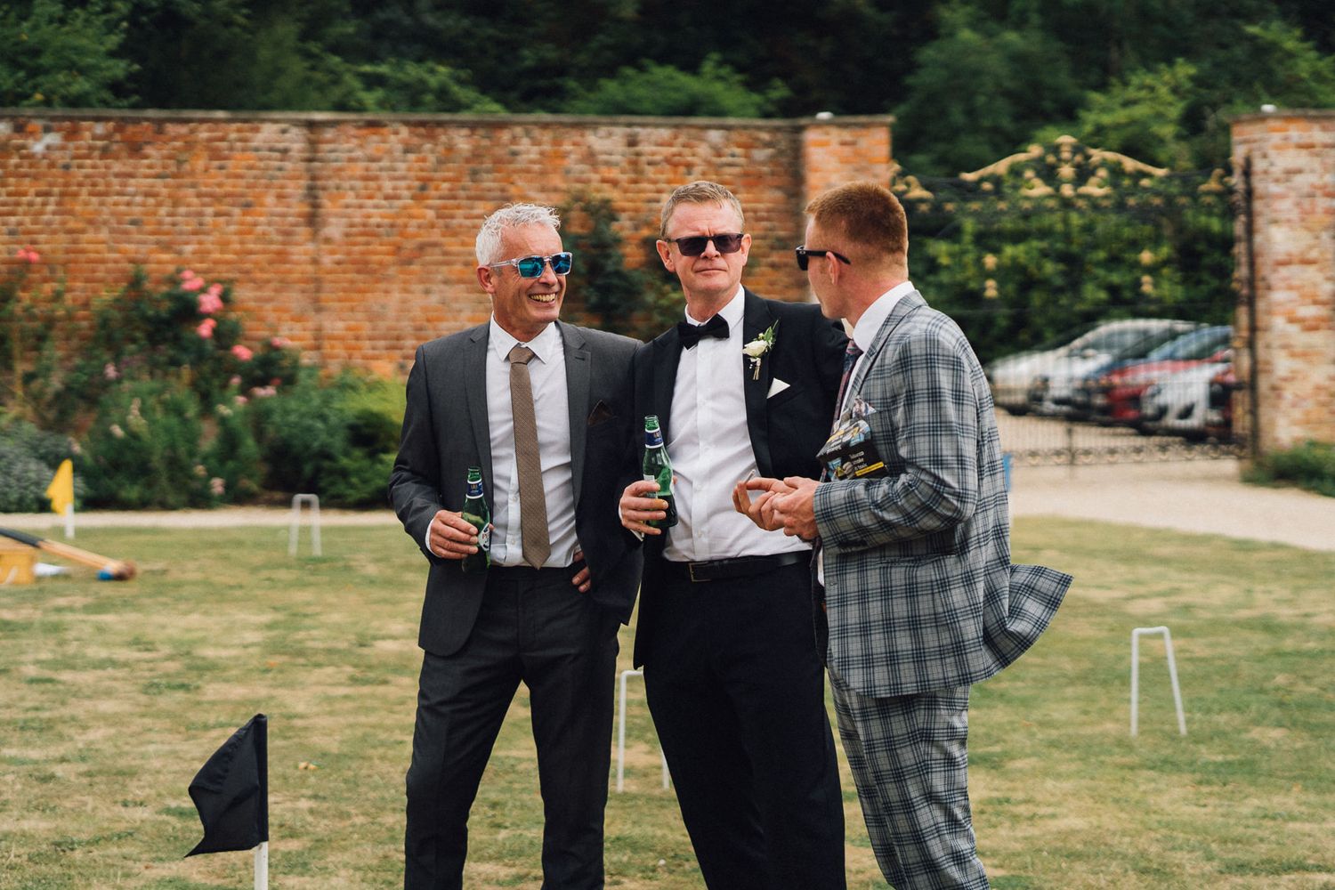 Wedding guests socialize on a lawn outside a brick building during a summer celebration.