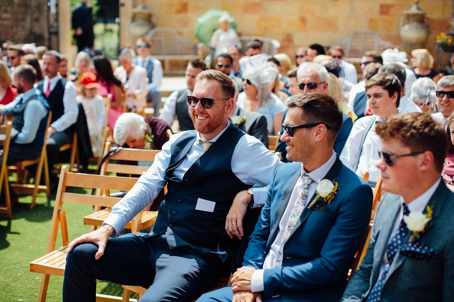 Wedding guests seated outdoors on wooden chairs during sunny garden ceremony.
