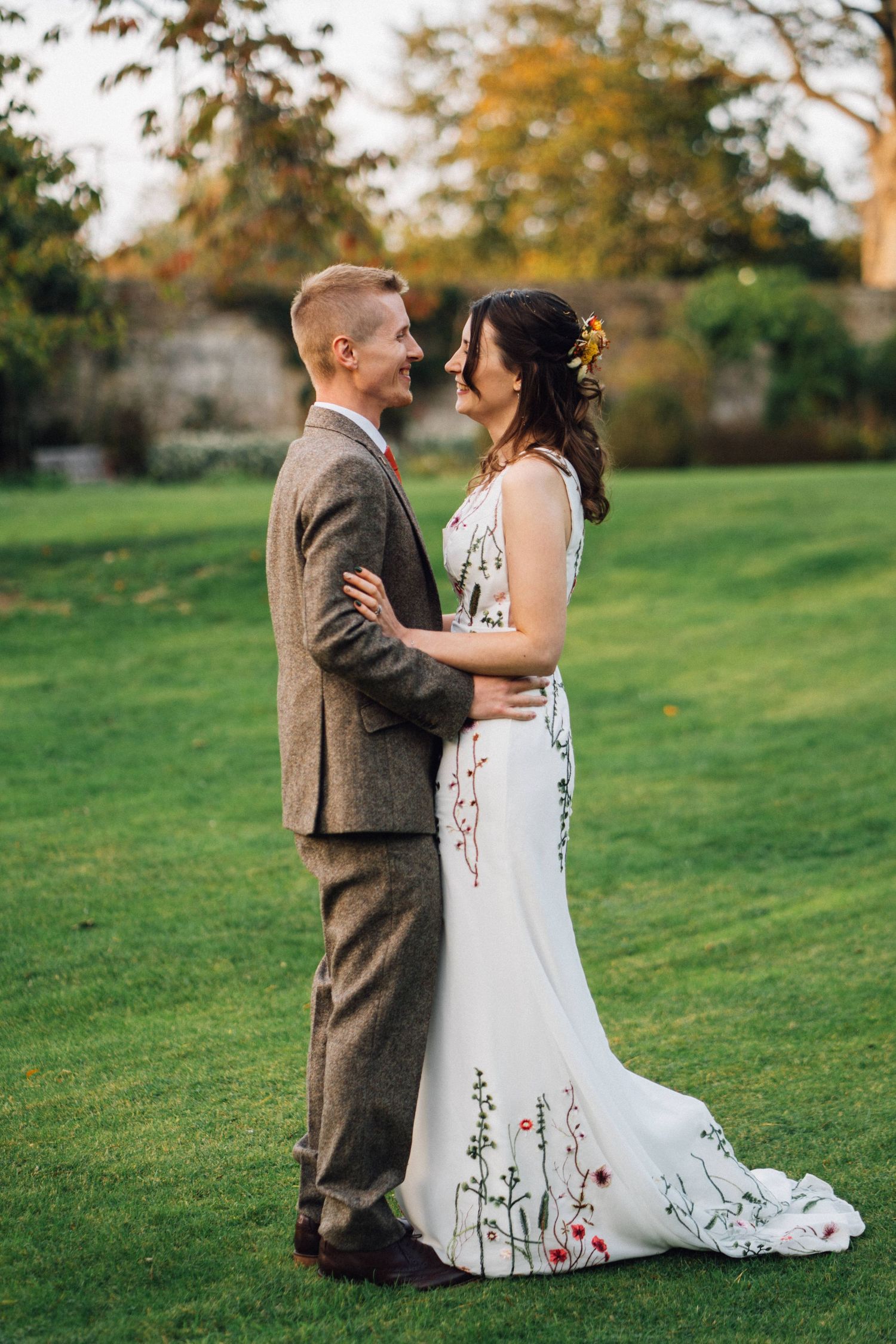 Couple embraces on green lawn wearing wedding attire with floral detailed dress.