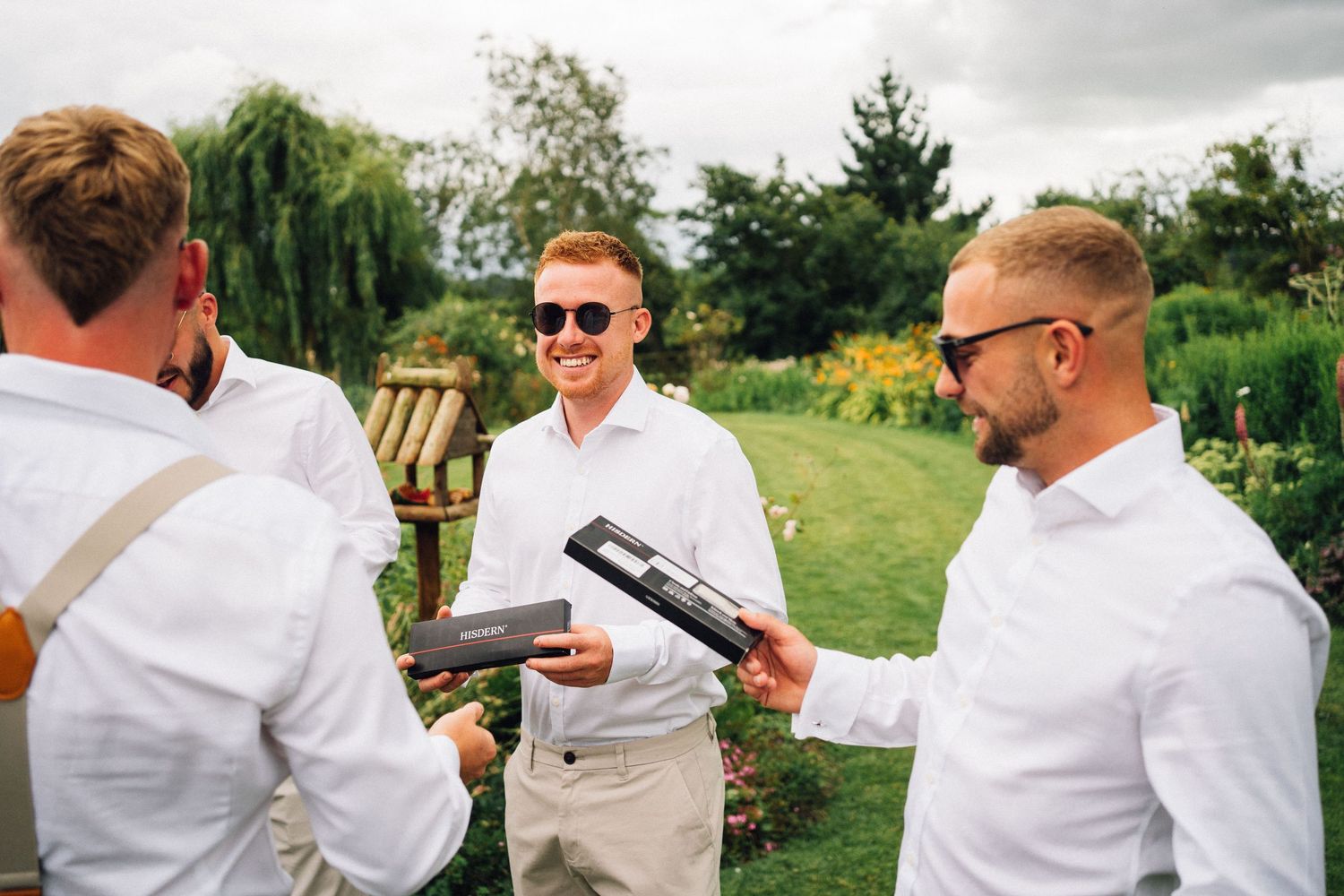 Three groomsmen in white dress shirts share drinks and conversation in an outdoor garden setting.