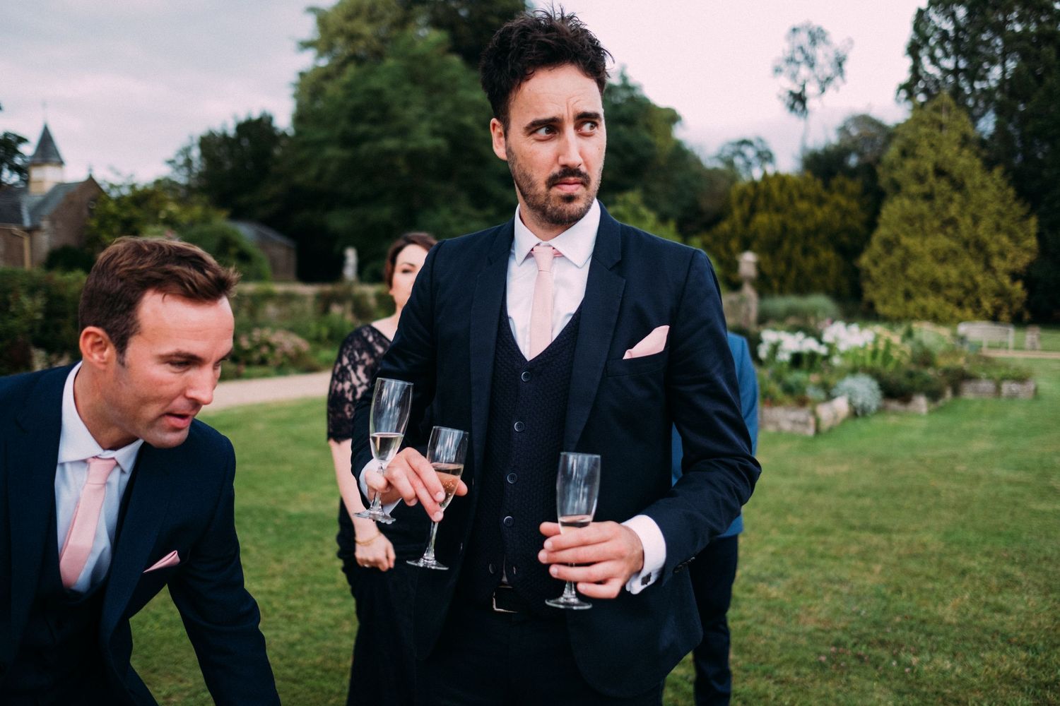 Groomsmen in navy suits socialize with drinks on the lawn of an elegant estate.