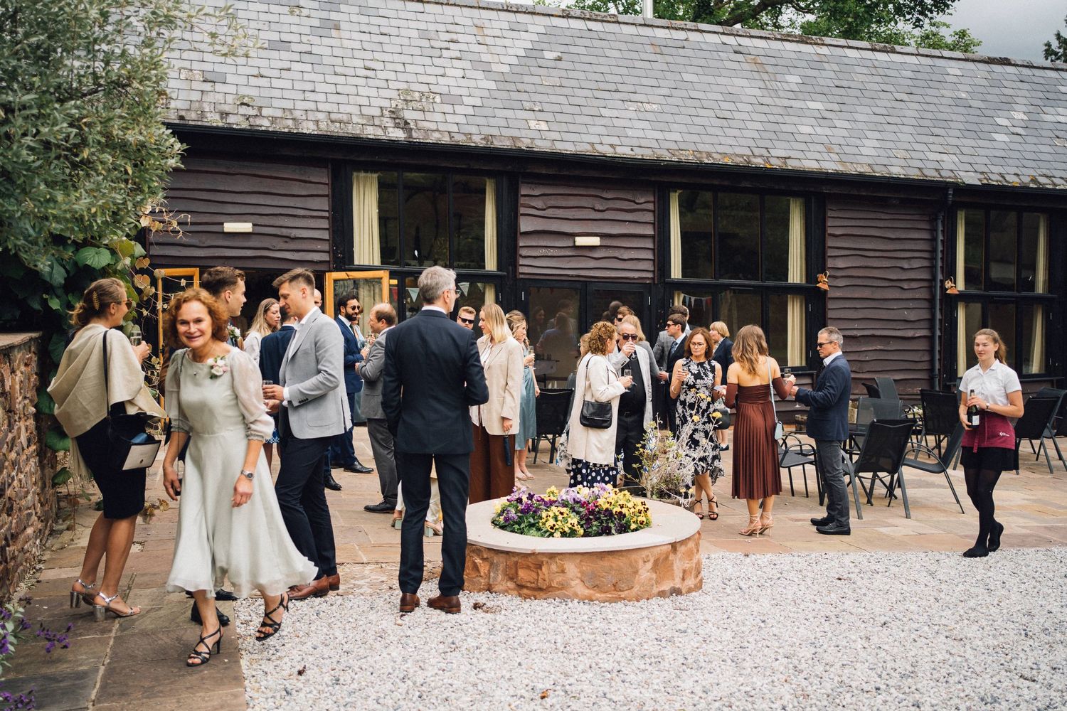 Wedding guests gathered outside a rustic wooden venue around a circular planter.