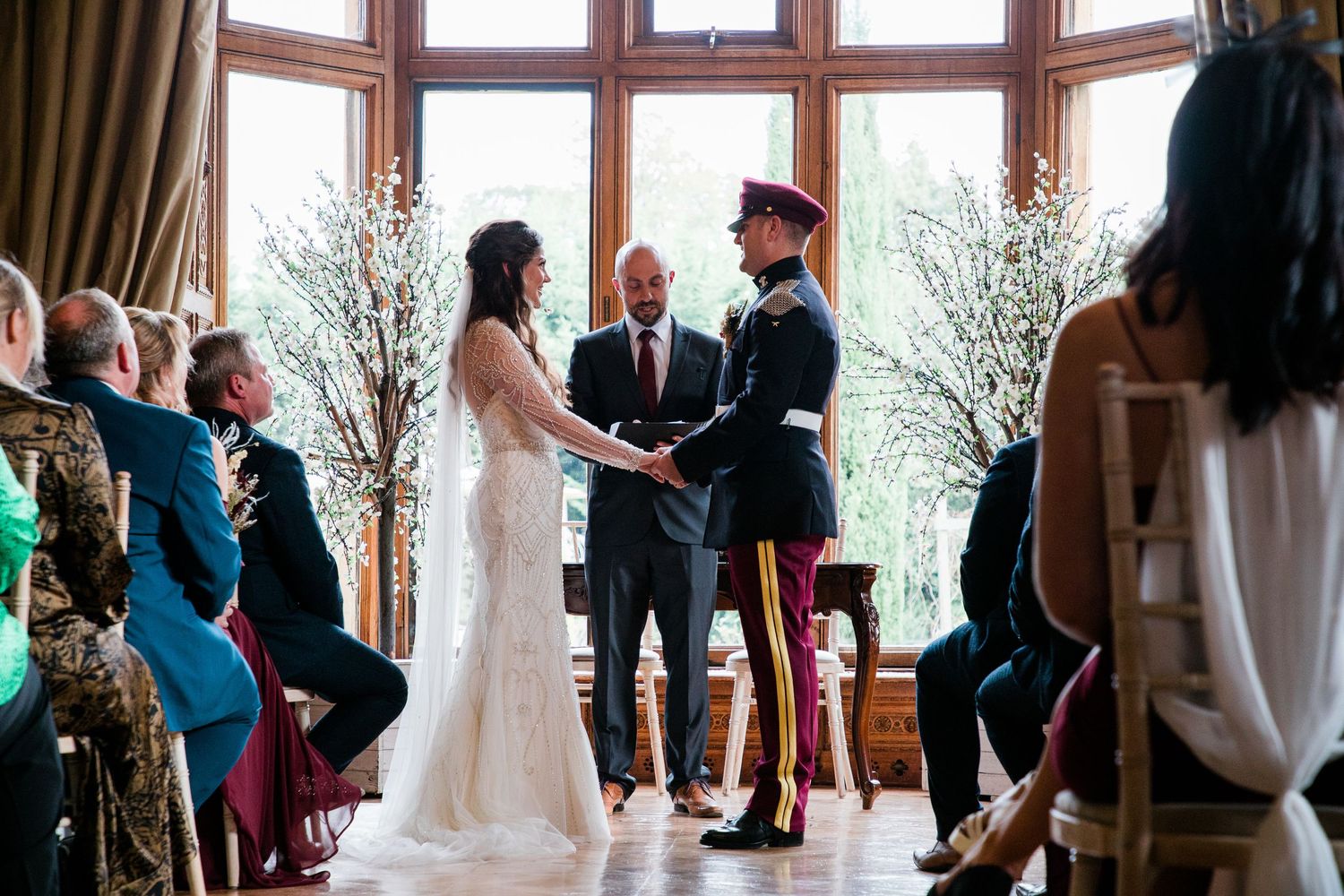 Wedding ceremony guests seated in rows inside an elegant room with large windows.