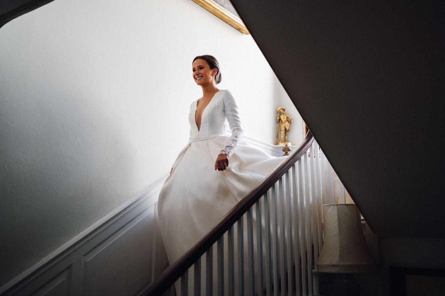 Bride in elegant long-sleeve white wedding gown descending a staircase, bathed in soft natural light.