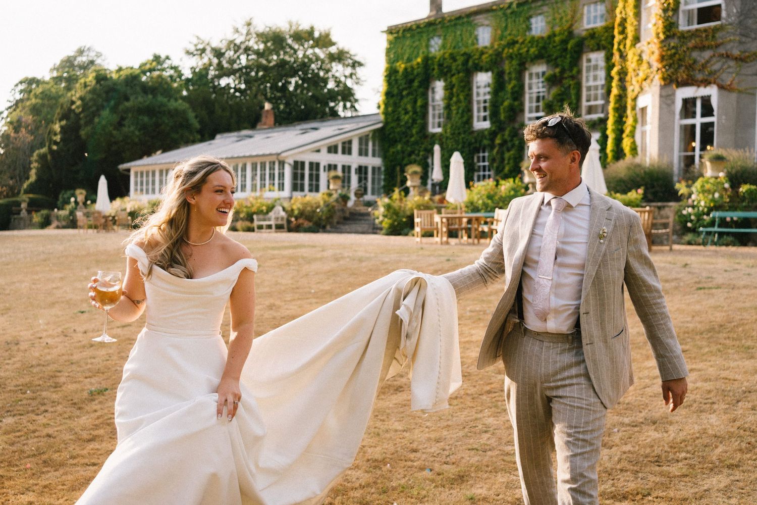 Bride and groom laughing outdoors at ivy-covered English country house wedding venue on sunny summer day.
