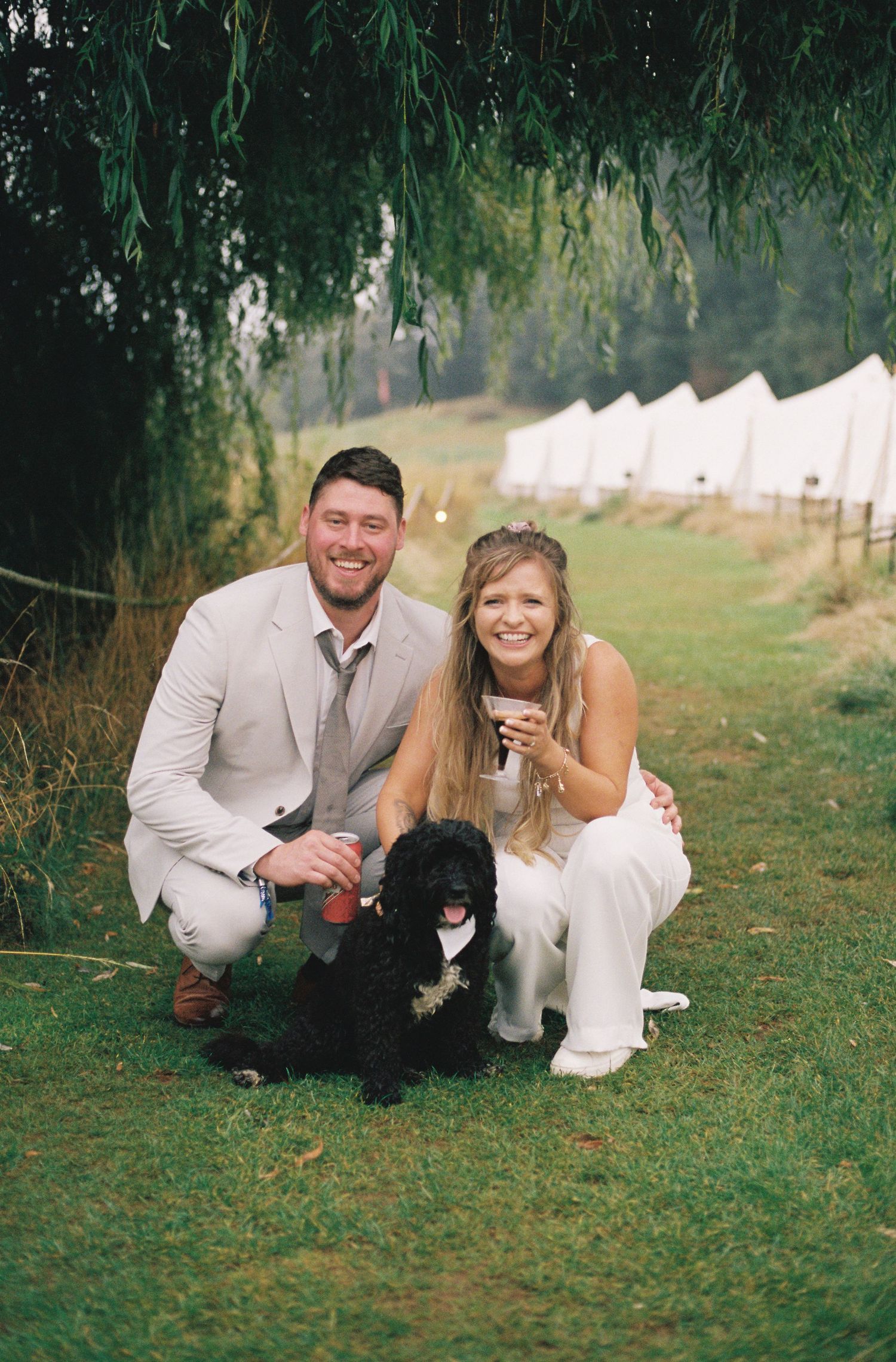 Smiling couple in wedding attire crouching with a black curly dog on green grass under willow trees with white tents.