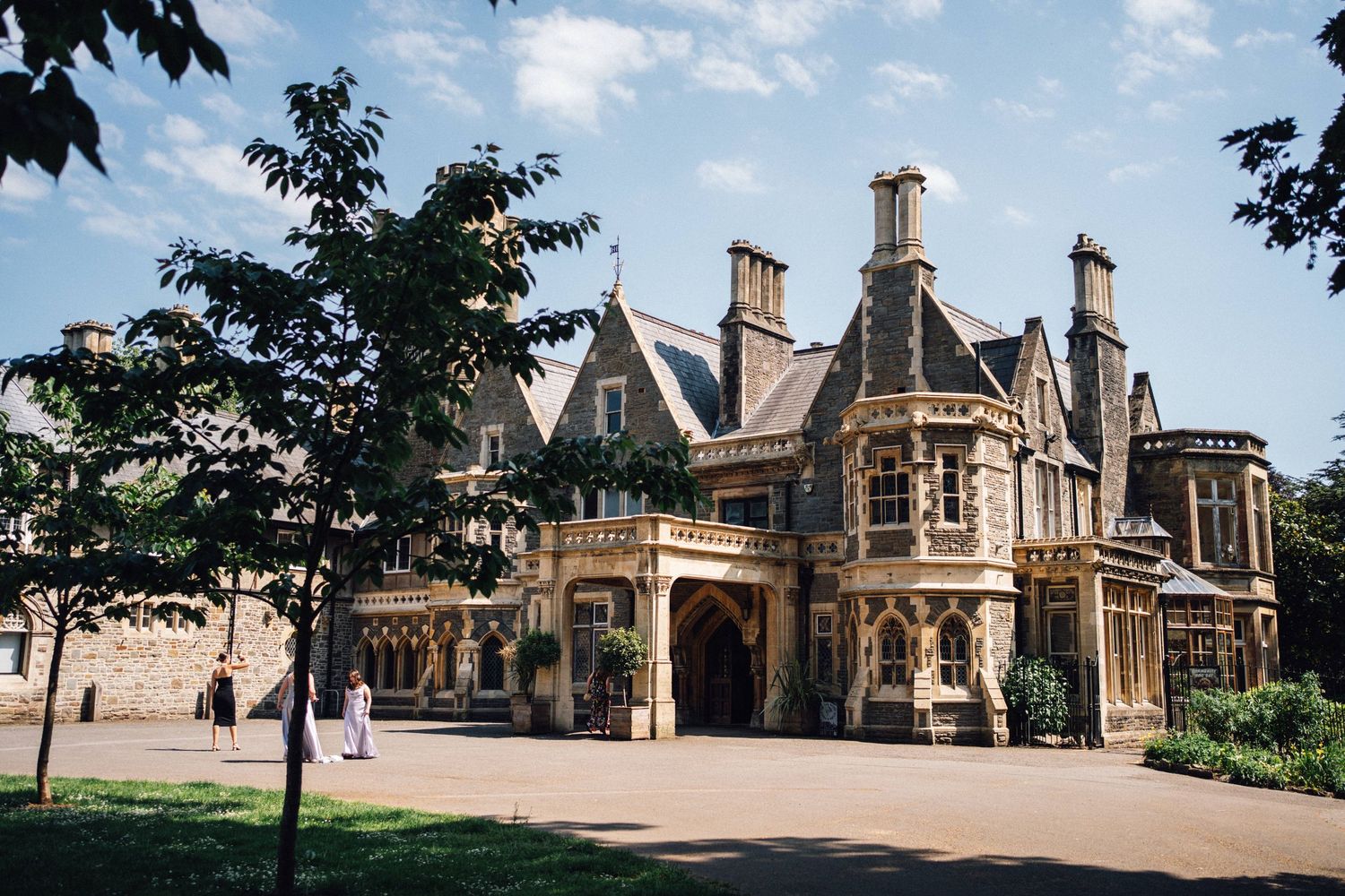 Grand Victorian manor house with ornate stone architecture, arched entrance, and manicured grounds under a blue sky.