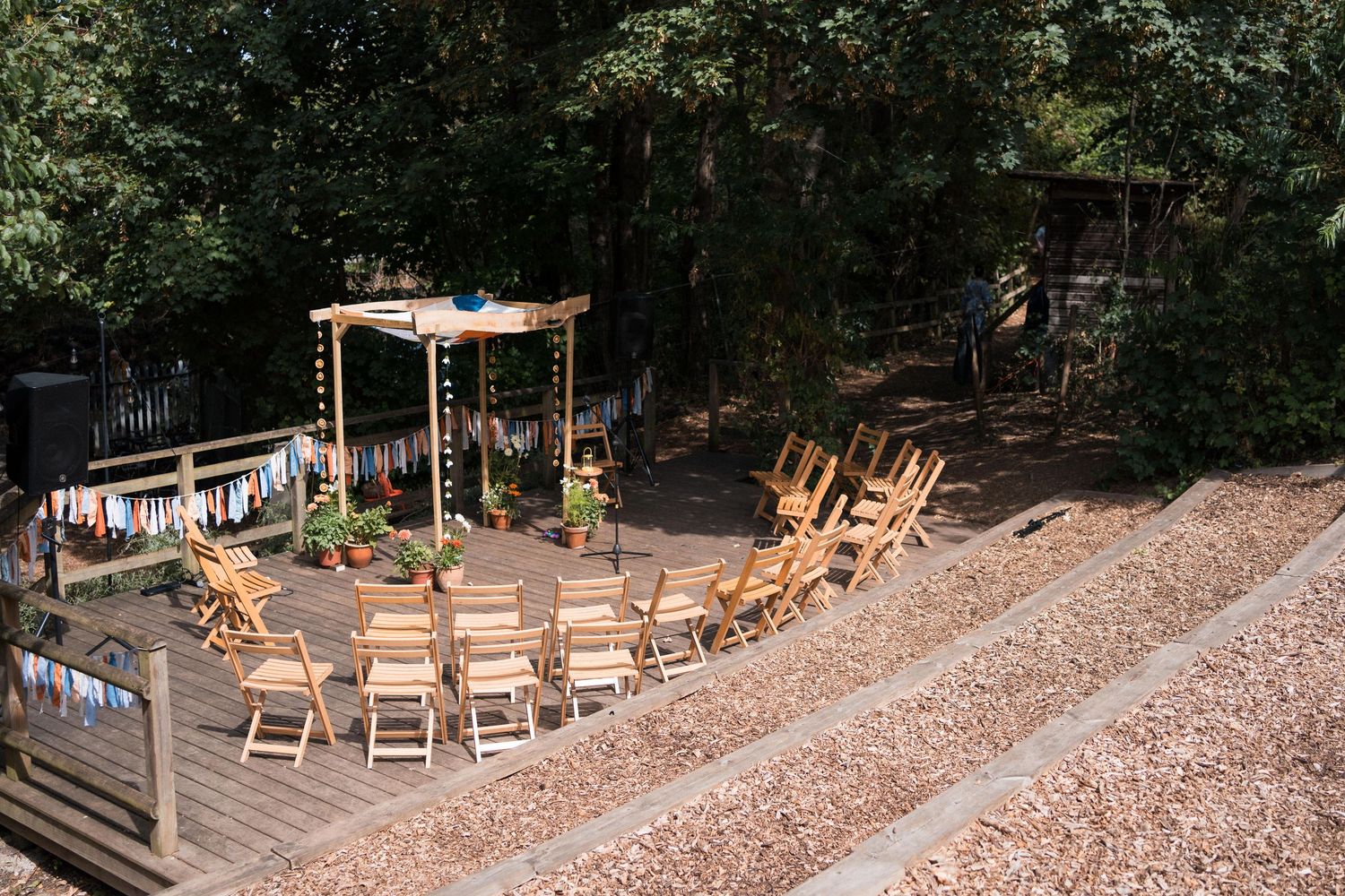 Outdoor wedding ceremony setup with wooden chairs arranged in a circle around a floral arch on a deck surrounded by trees.
