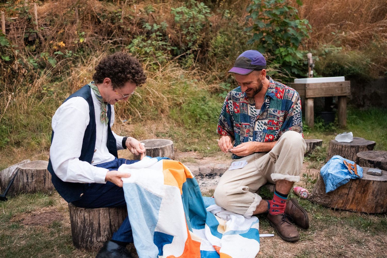 Two men sit outdoors on logs carefully sewing a colorful blue, orange, and white fabric together.