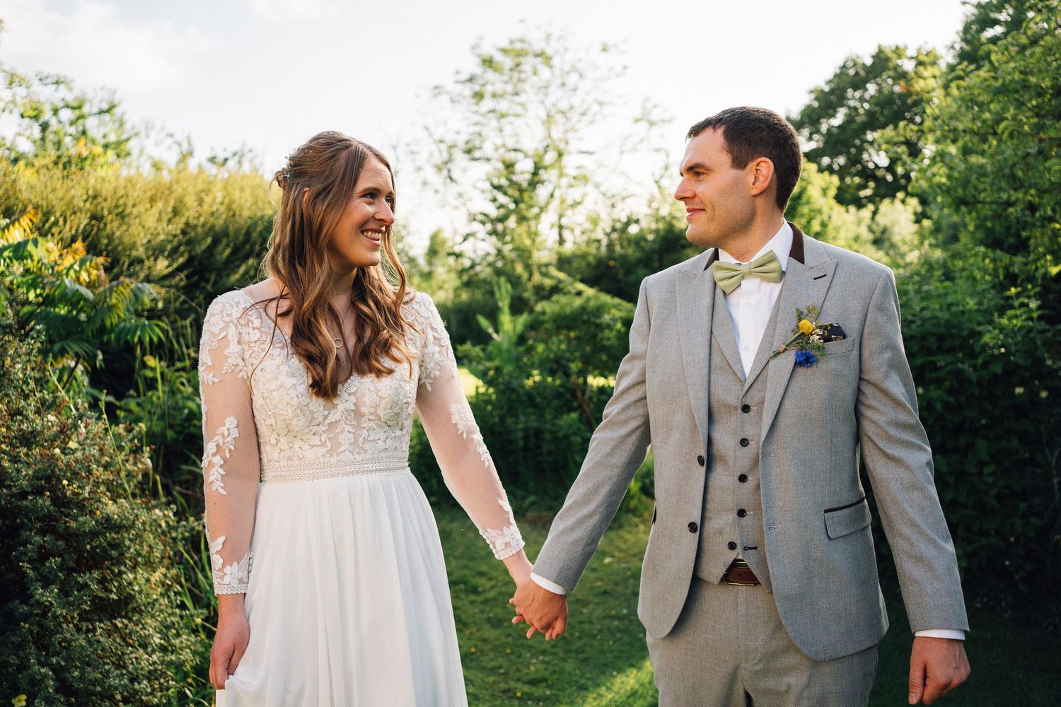 A smiling bride in a lace wedding dress holds hands with a groom in a gray suit outdoors.