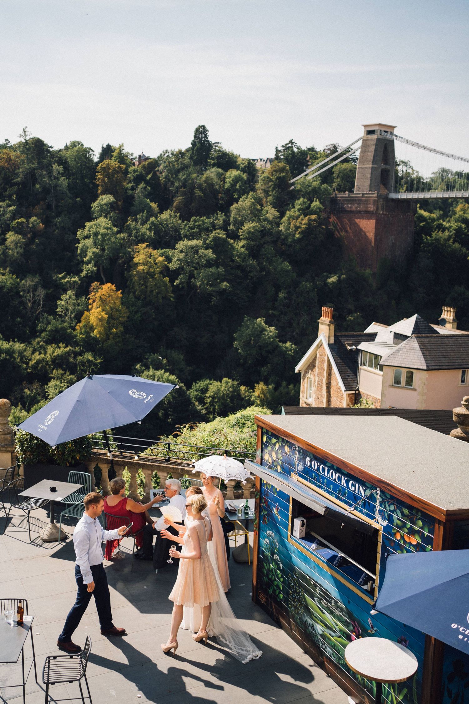 Outdoor wedding reception on a terrace with a gin bar, guests mingling, lush greenery, and a suspension bridge in the background.