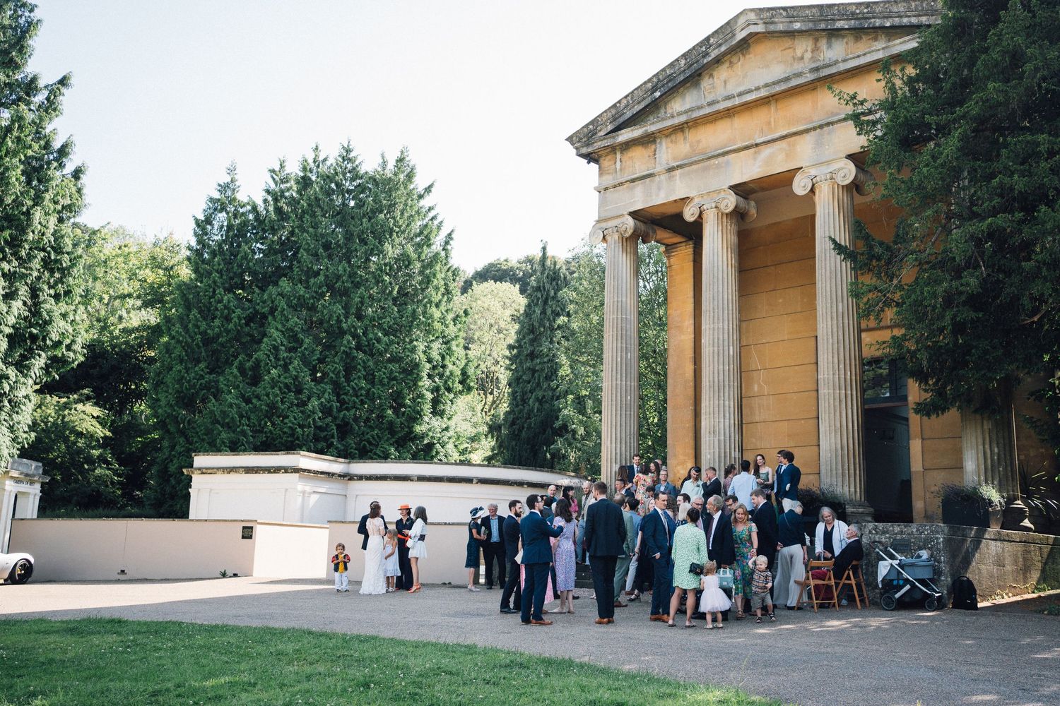 Elegant outdoor wedding reception at a neoclassical building with columns, guests mingling on terrace surrounded by lush green trees.