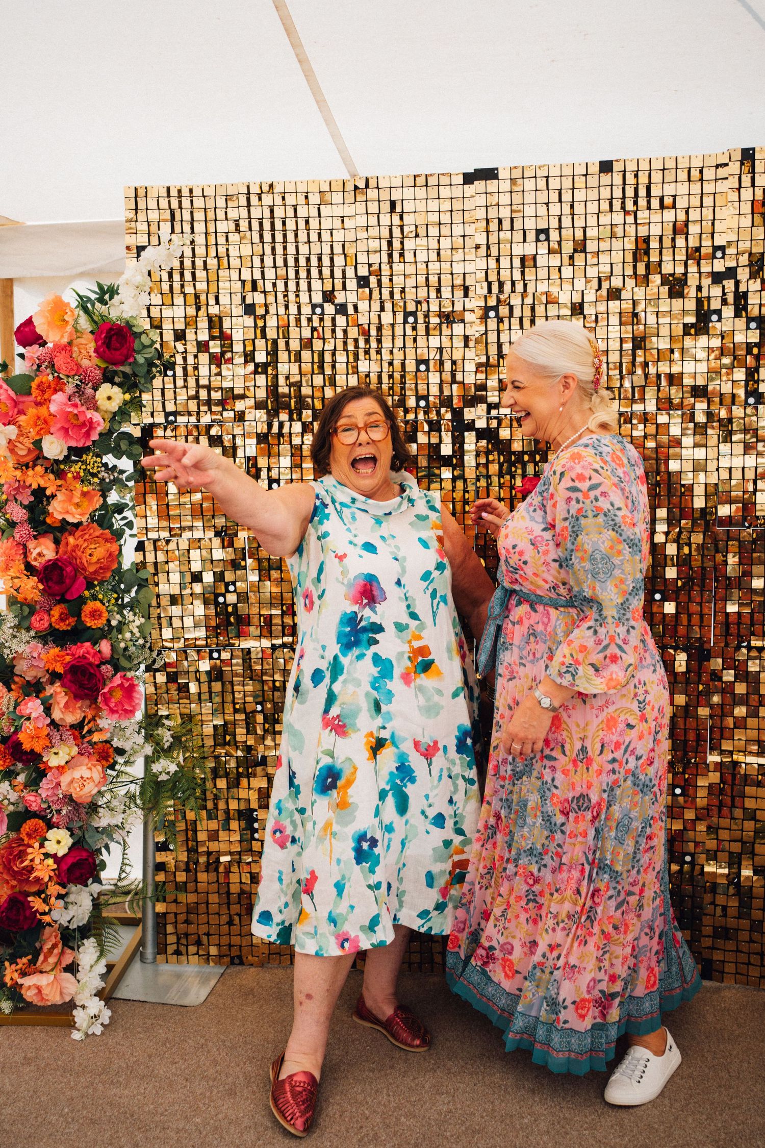 Two people in colorful floral dresses pose and dance in front of a sparkly gold sequin backdrop at a celebration.